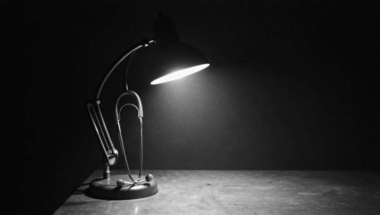 A desk lamp at night with a stethoscope hung over its arm, a black and white image about the practice that funds the build